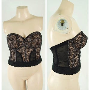 Pode incluir: Bustier de renda preta com decote em coração e barbatanas. O bustier apresenta um delicado padrão floral de renda sobre um forro nude, com painéis transparentes nas laterais e uma borda com vieiras na parte inferior. A peça é exibida em um manequim.
