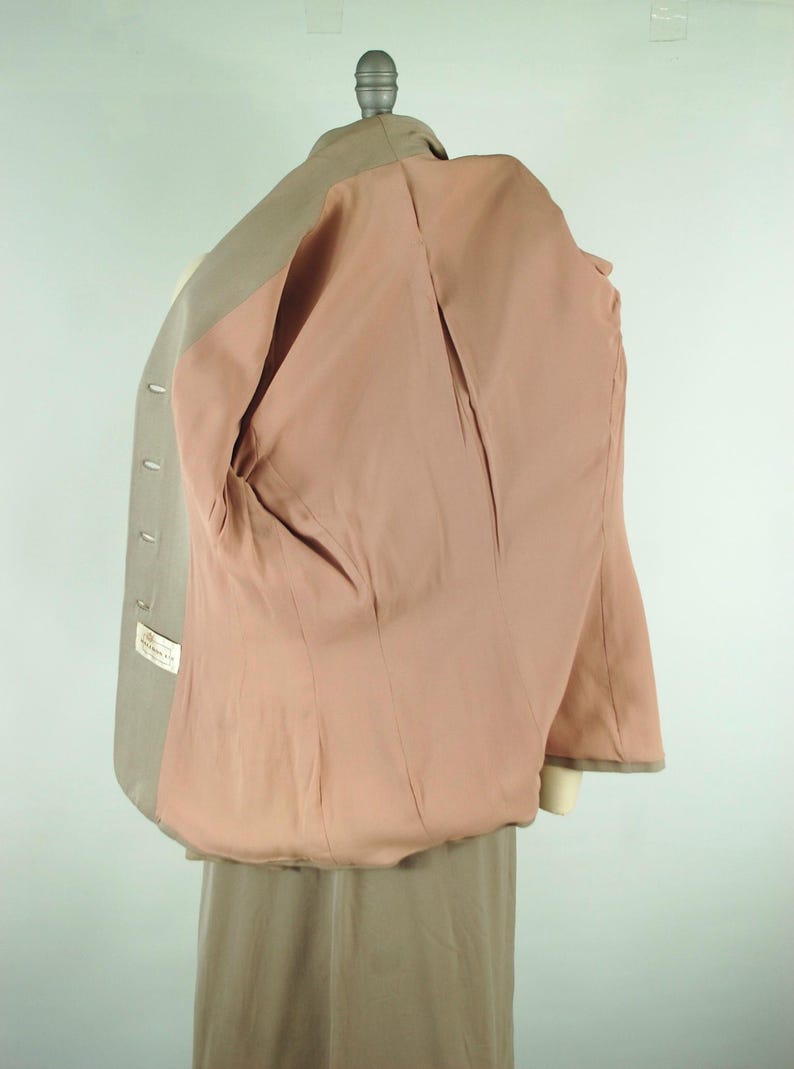 May include: A light tan blazer with a pink interior lining, displayed on a mannequin. The blazer has a classic design with a notched lapel and button closure. The interior lining is a solid pink color.