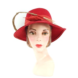 May include: A red hat with a wide brim, adorned with a red ribbon and gold accents. A decorative feather and a gold-colored accent are attached to the side. The hat is displayed on a mannequin head.
