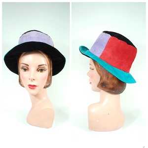 Op de afbeelding: Een kleurrijke hoed met een brede rand. De hoed heeft een zwarte rand, een paarse bovenkant, een rood zijpaneel en een turquoise accent. De hoed wordt gepresenteerd op een mannequinhoofd tegen een witte achtergrond.
