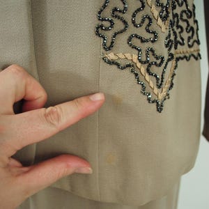 May include: A tan-colored suit jacket with decorative embellishments. The jacket features a detailed design on the front, with black beading and tan-colored trim. The fabric appears to be a smooth, woven material.