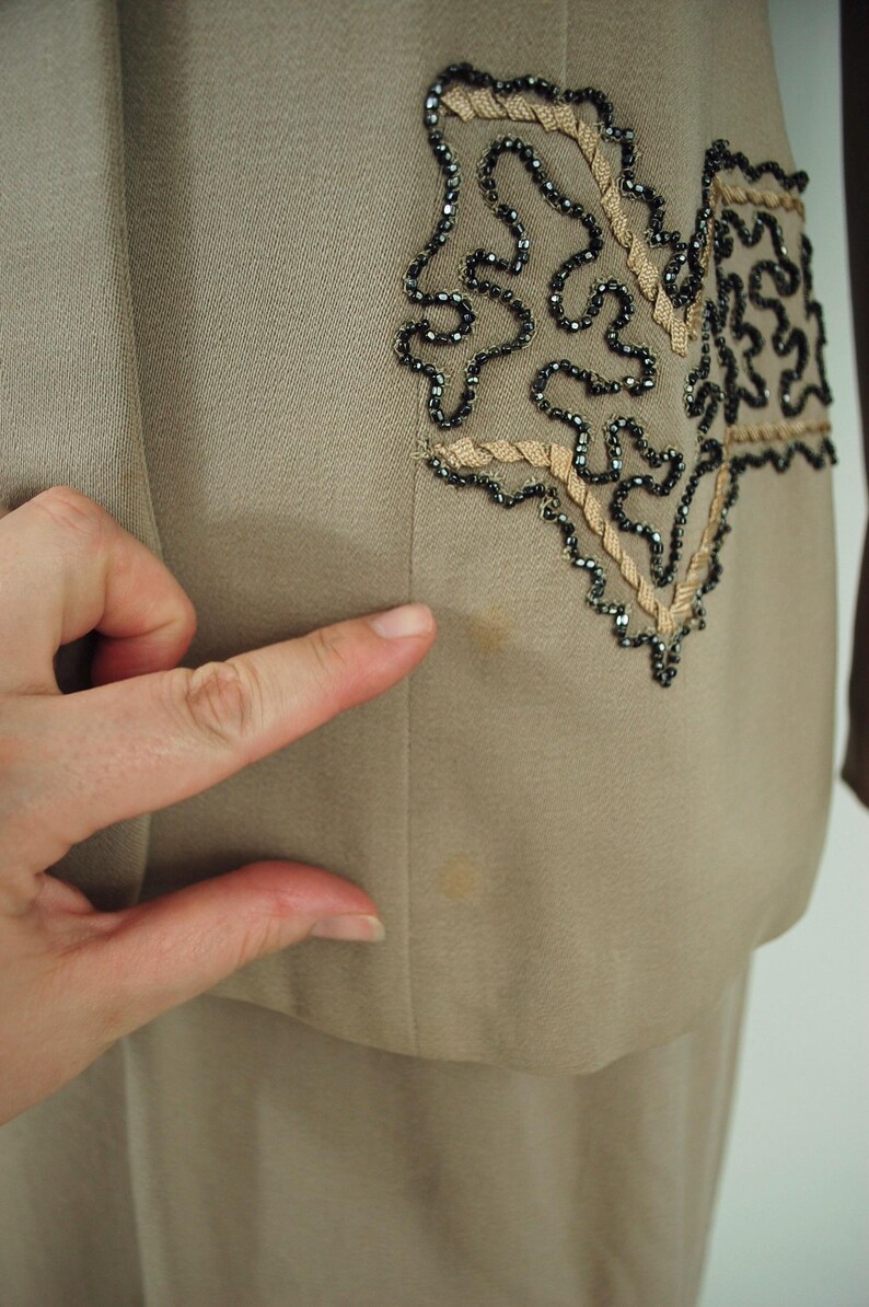 May include: A tan-colored suit jacket with decorative embellishments. The jacket features a detailed design on the front, with black beading and tan-colored trim. The fabric appears to be a smooth, woven material.