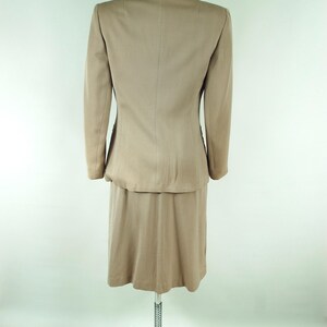 May include: A tan skirt suit displayed on a mannequin. The suit includes a blazer with long sleeves and a knee-length skirt. The fabric has a subtle sheen. The mannequin is on a wheeled stand against a white background.