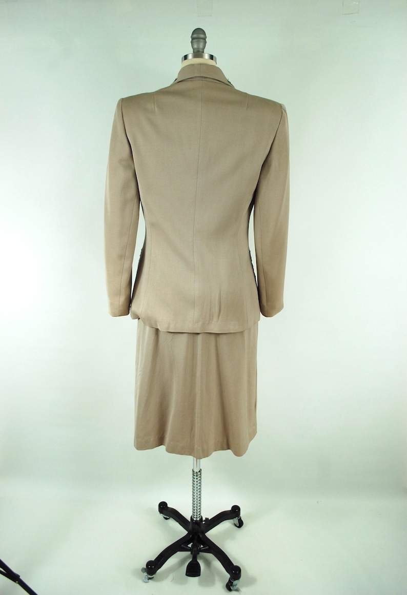 May include: A tan skirt suit displayed on a mannequin. The suit includes a blazer with long sleeves and a knee-length skirt. The fabric has a subtle sheen. The mannequin is on a wheeled stand against a white background.