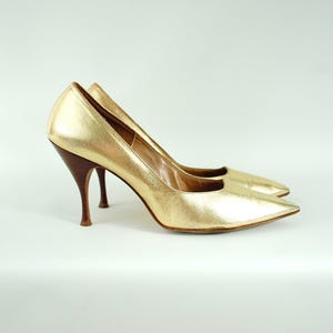 Könnte beinhalten: Ein Paar goldfarbene High Heels mit spitzer Zehenpartie. Die Absätze sind dunkelbraun. Die Schuhe stehen vor einem weißen Hintergrund.