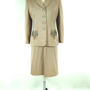 May include: A tan skirt suit displayed on a mannequin. The suit features a blazer with three buttons and decorative embroidery on the lapels and pockets. The skirt is knee-length. The suit is made of a smooth fabric.