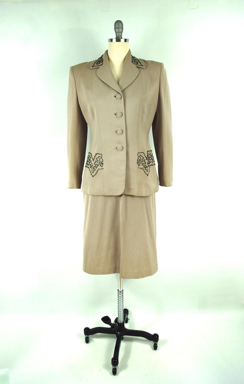 May include: A tan skirt suit displayed on a mannequin. The suit features a blazer with three buttons and decorative embroidery on the lapels and pockets. The skirt is knee-length. The suit is made of a smooth fabric.