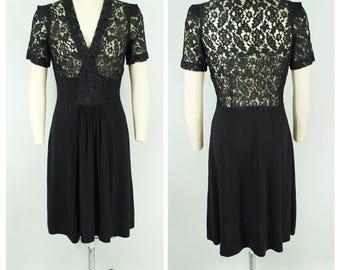 May include: A black vintage dress with a V-neckline and short sleeves. The bodice features a lace overlay, and the skirt is a simple, flowing design. The dress is displayed on a mannequin, showcasing the front and back.