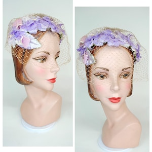 May include: A vintage hat with a floral design and a veil. The hat features a crown of lavender and white fabric flowers. A fine, beige netting veil drapes over the face. The hat is displayed on a mannequin head with red lipstick.