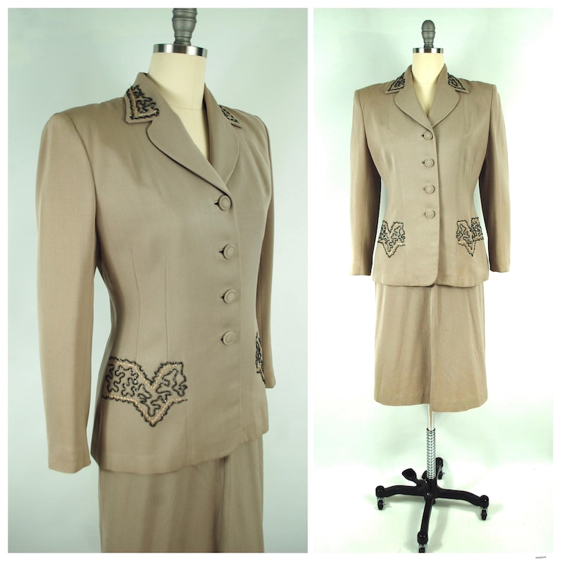 May include: A tan vintage skirt suit displayed on a mannequin. The suit features a tailored blazer with a notched collar, button closure, and decorative embroidery. The matching skirt is knee-length, creating a classic, professional ensemble.
