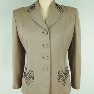 May include: A tan two-piece suit with a blazer and skirt. The blazer features black and gold embroidered detailing on the lapels and pockets, with matching buttons down the front. The suit is displayed on a mannequin.