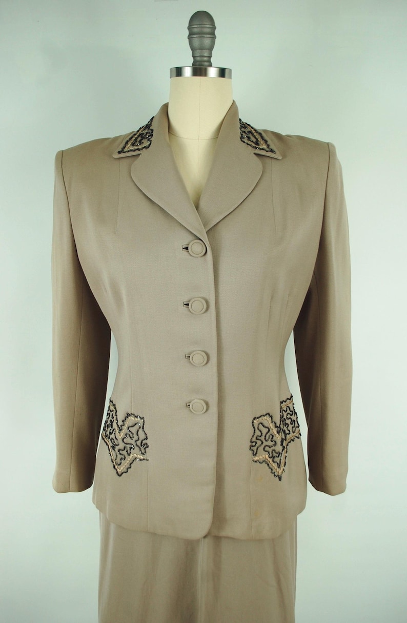 May include: A tan two-piece suit with a blazer and skirt. The blazer features black and gold embroidered detailing on the lapels and pockets, with matching buttons down the front. The suit is displayed on a mannequin.