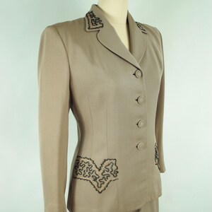 May include: A tan two-piece suit displayed on a mannequin. The jacket features a notched collar, three buttons, and decorative black and gold embroidery on the lapels and pockets. The skirt is a matching tan color.