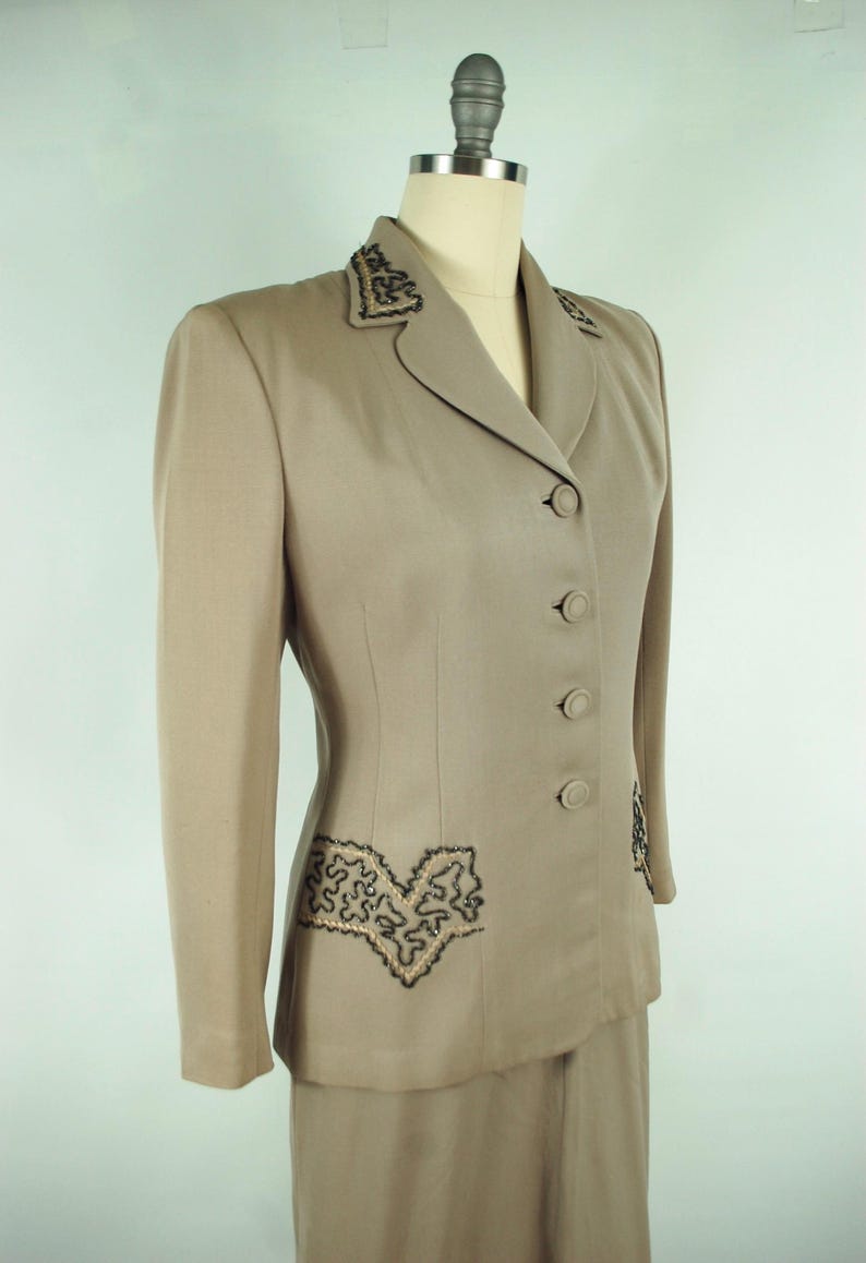 May include: A tan two-piece suit displayed on a mannequin. The jacket features a notched collar, three buttons, and decorative black and gold embroidery on the lapels and pockets. The skirt is a matching tan color.