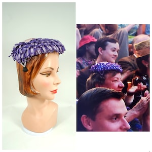 Puede incluir: Un sombrero vintage morado y blanco con un diseño floral. El sombrero se exhibe en una cabeza de maniquí. El sombrero tiene un ribete decorativo y una pequeña borla negra. Un accesorio único.