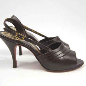 Puede incluir: Un par de zapatos de tacón alto de color marrón oscuro con punta abierta, correa en el tobillo y hebilla dorada. Los tacones de aguja tienen suelas rojas. Los zapatos son de cuero. Los tacones miden unos 10 cm.