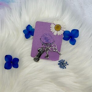 May include: A silver charm keyring with a ball of yarn, scissors, and a ring. The keyring is on a purple background with dried flowers.