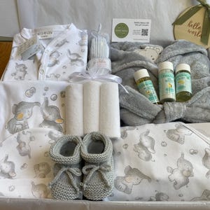 May include: A baby gift set containing a white babygrow, a grey robe, baby wash, washcloths, and grey knitted booties. The items are arranged on a white blanket with elephant illustrations. The gift set is presented in a white tray.