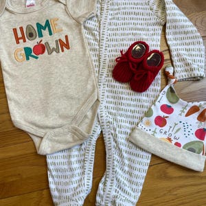 May include: A baby clothing set including a beige onesie with "HOME GROWN" text, a patterned footed sleeper, red knit booties, and a matching hat with fruit and vegetable illustrations. The set is designed for newborns.