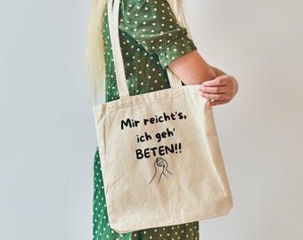 May include: A white tote bag with black text that reads "Mir reicht's, ich geh' BETEN!!" and a simple black line drawing of two hands clasped together. The bag is being held by a person wearing a green polka dot dress.