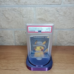Puede incluir: Una carta de comercio de Pokemon clasificada con Pikachu usando un sombrero de fieltro gris. La carta está en una caja protectora y se muestra en un soporte morado. La carta está clasificada GEM MT 10 por PSA.