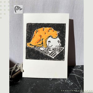 May include: A whimsical art print featuring a stylized orange cat atop a white figure with a smiling face, set against a black background. The cat and figure are on a striped mat. The artwork is on a white rectangular paper.