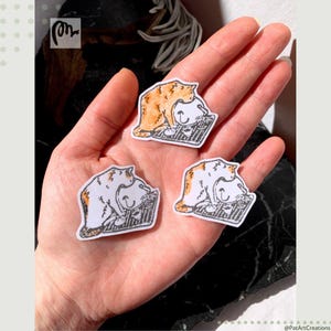 May include: Three cat stickers are held in a hand. Each sticker features a white cat with orange tabby markings, curled up on a black and grey striped surface. The stickers are hand-drawn with a whimsical style.