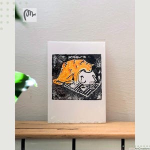 May include: A rectangular art print featuring a linocut-style illustration of an orange tabby cat leaning over a white teapot. The cat is positioned on a black textured background with a small white cup and a striped mat visible beneath the teapot.