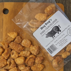 May include: A clear plastic bag of Day Day's Pork Cracklings, fried-out pork fat with attached skin, sits on a wooden cutting board. The bag's label features a pig illustration and ingredient list. Scattered around the bag are pieces of the cracklings.