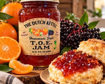 May include: A glass jar of The Dutch Kettle T.O.E. Jam with a gold lid, featuring a label with text, orange and elderberry illustrations, and a net weight of 19 ounces. A biscuit topped with the red jam sits on a plate next to the jar.