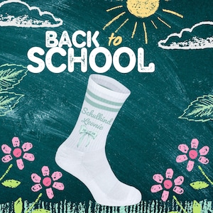 Könnte beinhalten: Weiße Socke mit mintgrünen Streifen und dem Text "Schulkind Leonie 2026" auf einem Tafelhintergrund mit den Worten "Back to School". Das Bild zeigt auch gezeichnete Blumen, Wolken und eine Sonne.