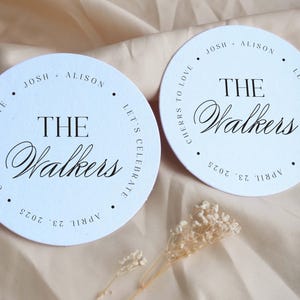 K&ouml;nnte beinhalten: Zwei wei&szlig;e runde Untersetzer mit schwarzem Text. Der Text lautet "Cheers to love, Josh & Alison, The Walkers, Let's Celebrate, April 23, 2025".