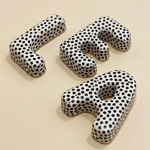 May include: Three ceramic letters, J, E, and R, are arranged on a neutral surface. Each letter is white with a pattern of black polka dots. The letters are three-dimensional and have a smooth, glossy finish.
