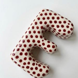 May include: A three-dimensional letter E with a glossy off-white finish and a repeating pattern of small, dark red polka dots. The letter has rounded edges and a smooth, curved surface, creating a soft, rounded appearance across its entire form.