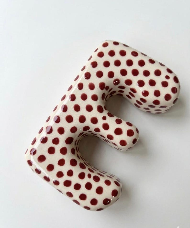 May include: A three-dimensional letter E with a glossy off-white finish and a repeating pattern of small, dark red polka dots. The letter has rounded edges and a smooth, curved surface, creating a soft, rounded appearance across its entire form.