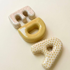 Op de afbeelding: Drie keramische letters: "E", "D" en "P". De "E" is gespikkeld beige, de "D" is effen geel en de "P" is wit met gele stippen. De letters zijn diagonaal gerangschikt op een crèmekleurig oppervlak.