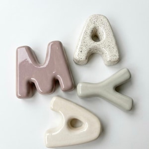 May include: Ceramic letters spelling out the word "MAY" in a playful arrangement. The "M" is a muted mauve, the "A" is speckled off-white, and the "Y" is a soft green. The second "A" is a creamy off-white. These decorative letters are likely for home decor.