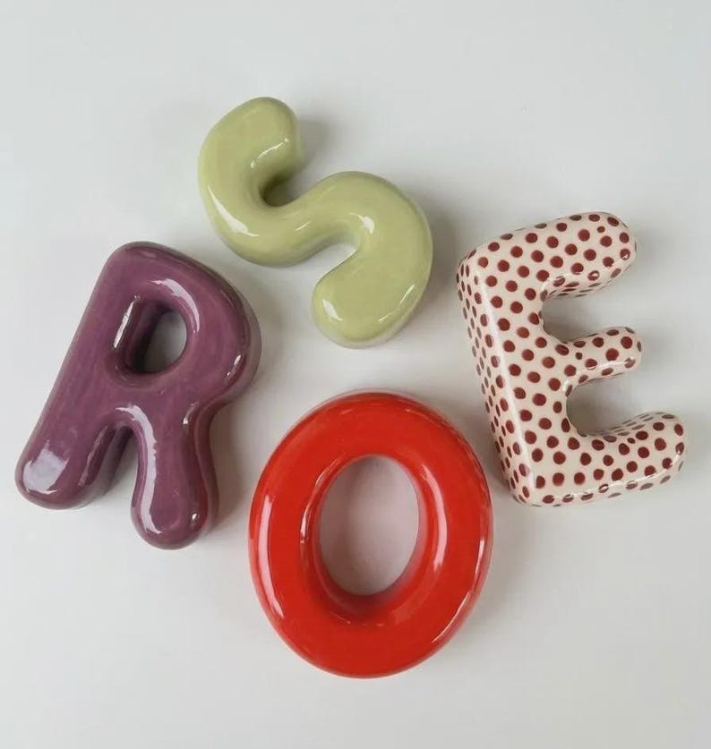 May include: Four ceramic letters arranged to spell the word ROSE. The letter R is solid purple, the letter O is solid red, the letter S is solid light green, and the letter E is white with a pattern of small brown polka dots. Each letter is glossy.