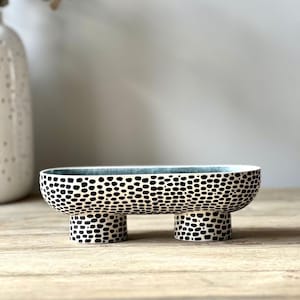 Może przedstawiać: Biała ceramiczna miska z czarno-białym wzorem w kropki. Miska ma trzy nóżki i stoi na drewnianej powierzchni.