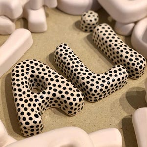 May include: A collection of three-dimensional ceramic letters, including the letters A and L, featuring a glossy white finish with black polka dots. Surrounding these are several plain, matte, off-white ceramic letters in various shapes.