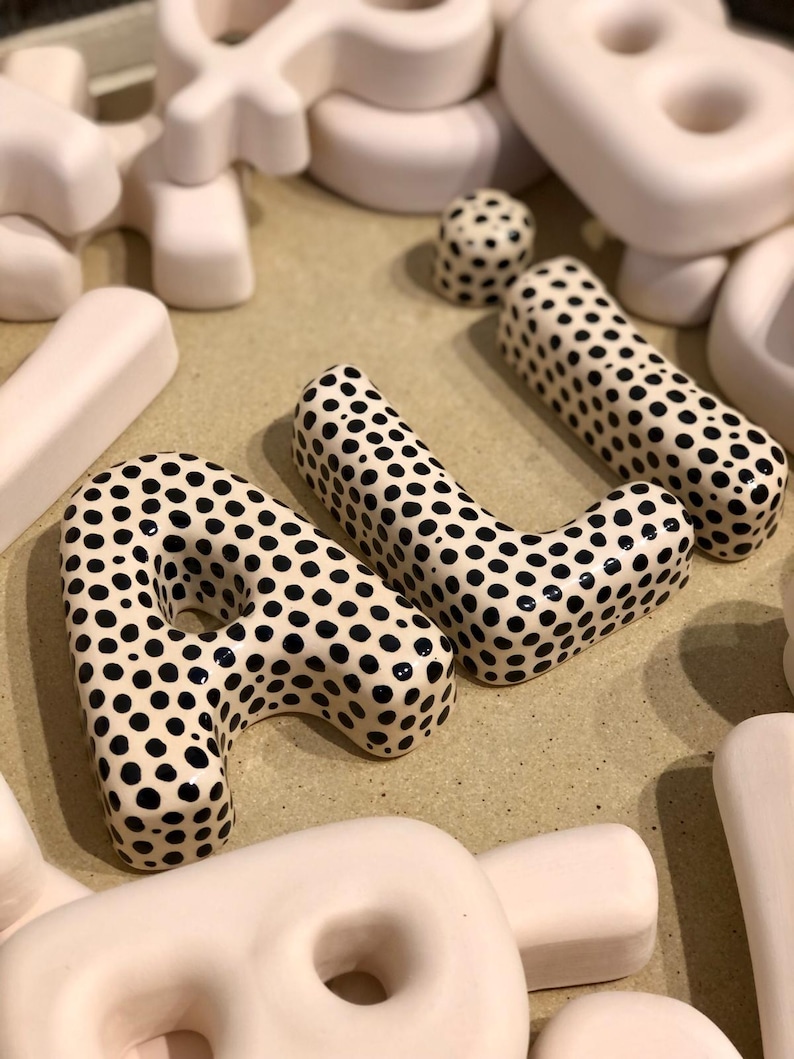 May include: A collection of three-dimensional ceramic letters, including the letters A and L, featuring a glossy white finish with black polka dots. Surrounding these are several plain, matte, off-white ceramic letters in various shapes.