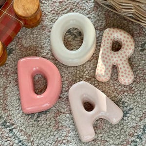 May include: Four ceramic letters spelling DORA. The letter D is solid pink, the letter O is solid white, the letter R is white with pink polka dots, and the letter A is a light, speckled pink. Each letter has a rounded, puffy, three-dimensional shape.