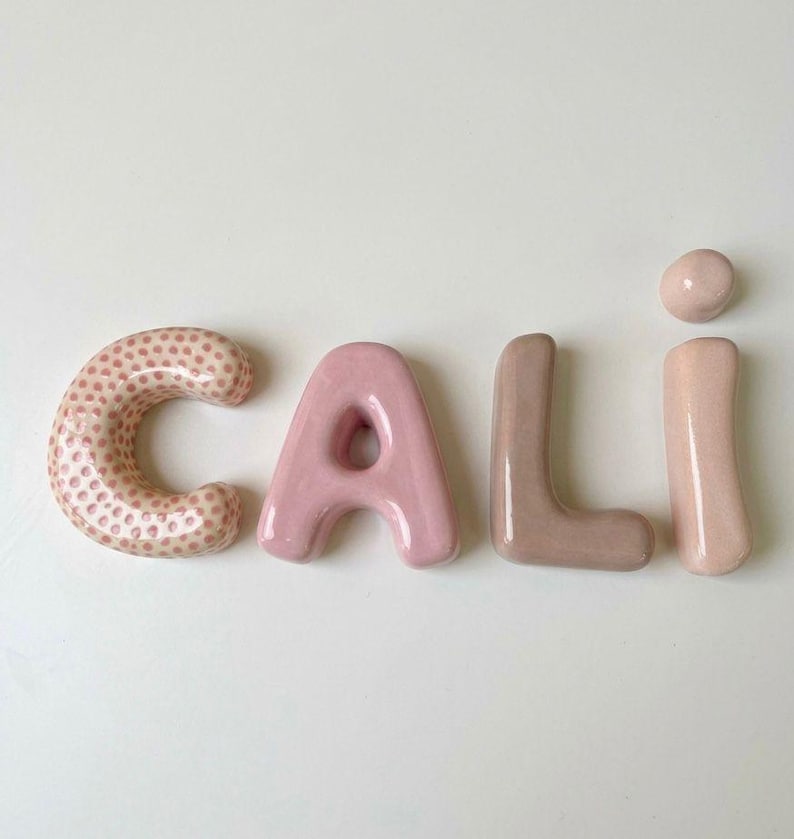 May include: Four individual ceramic letters spelling the name CALI. The letter C is light pink with a darker pink polka dot pattern. The letter A is a solid light pink. The letter L is a solid muted mauve. The letter I is a solid light pink.