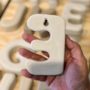 May include: A matte off-white ceramic letter E with rounded edges and a smooth finish. The back of the letter features a built-in keyhole slot for wall mounting, allowing it to be hung securely. The surface is uniform in color and texture.