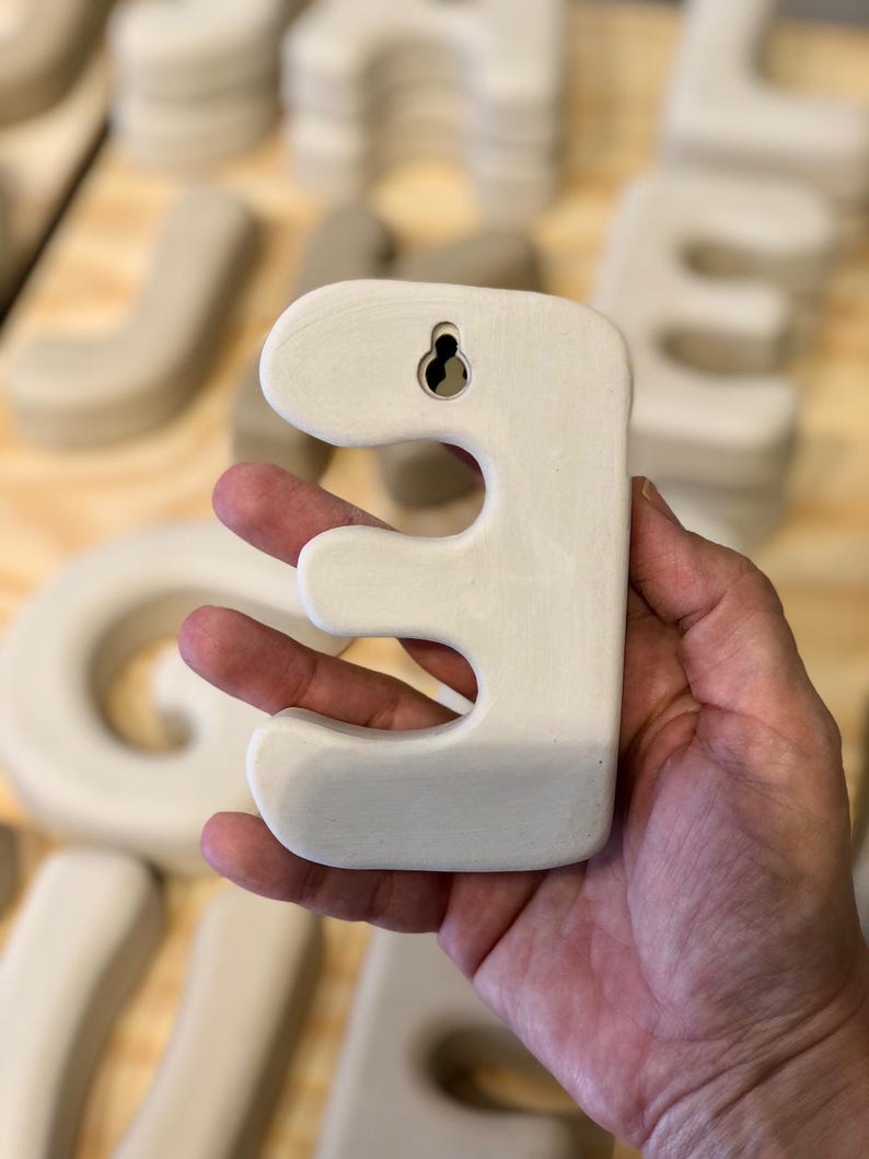 May include: A matte off-white ceramic letter E with rounded edges and a smooth finish. The back of the letter features a built-in keyhole slot for wall mounting, allowing it to be hung securely. The surface is uniform in color and texture.