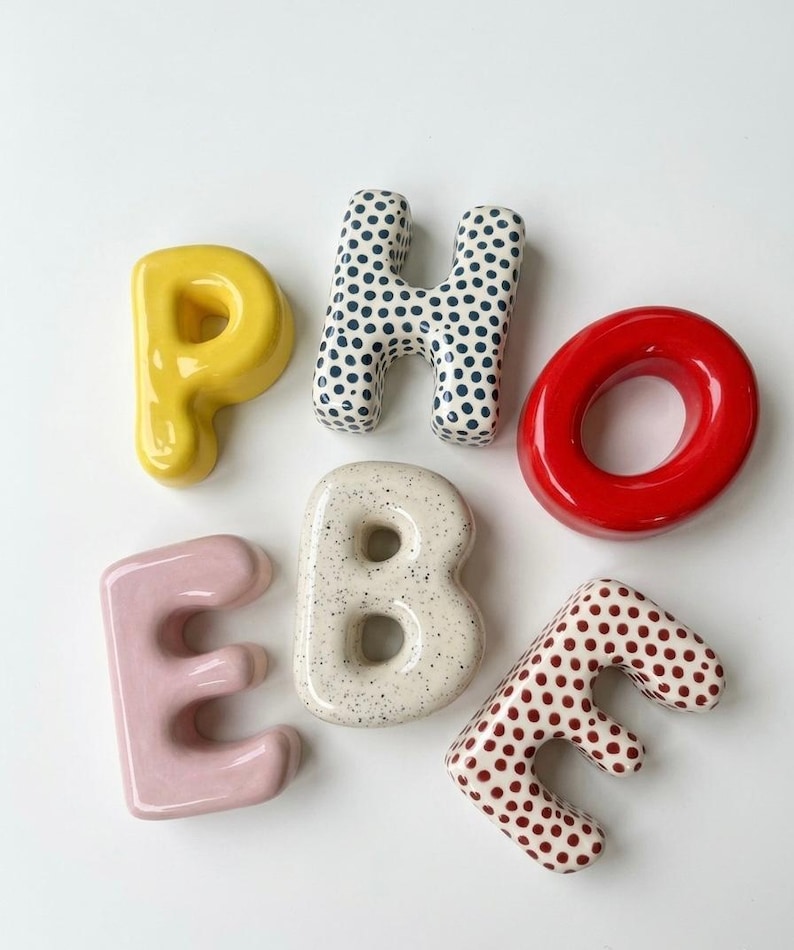 May include: A collection of six ceramic letters spelling PHOEBE. The P is solid yellow, the H has blue polka dots, the O is solid red, the E is solid light pink, the B is speckled cream, and the final E has red polka dots on a white base.