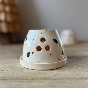 Könnte beinhalten: Cremefarbener Kerzenhalter aus Keramik mit Untertasse. Der Halter hat drei kreisförmige Öffnungen und ist mit grünen Weihnachtsbäumen und roten Punkten verziert. Im Hintergrund ist ein Weihnachtsbaum aus Holz zu sehen.