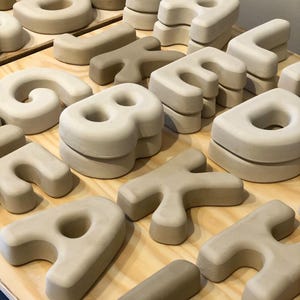 May include: A collection of three-dimensional, matte-finish ceramic letters in various shades of beige and off-white. The letters are shaped with rounded edges and thick, blocky forms, arranged in multiple stacks across a light-colored wooden surface.