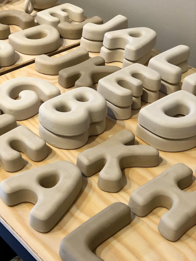 May include: A collection of three-dimensional, matte-finish ceramic letters in various shades of beige and off-white. The letters are shaped with rounded edges and thick, blocky forms, arranged in multiple stacks across a light-colored wooden surface.