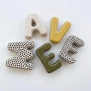 May include: Five ceramic letters spelling the word AVE. The letter A is speckled off-white, the V is solid mustard yellow, the E is solid olive green, and two letters M are white with black polka dots. Each letter has a smooth, rounded finish.